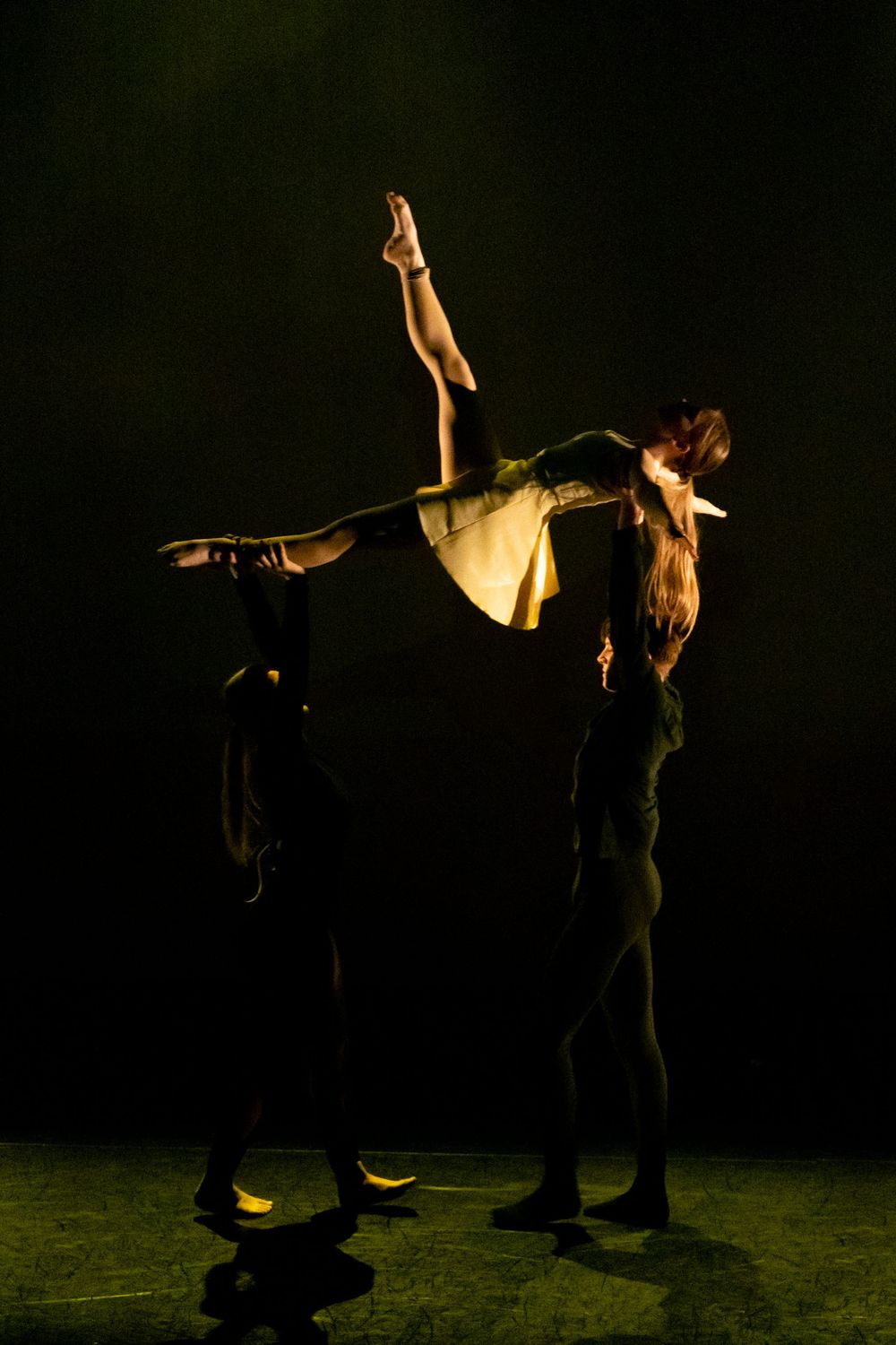 Two dancers dressed in black hold a ballerina in a white dress above their heads under strong yellow lighting