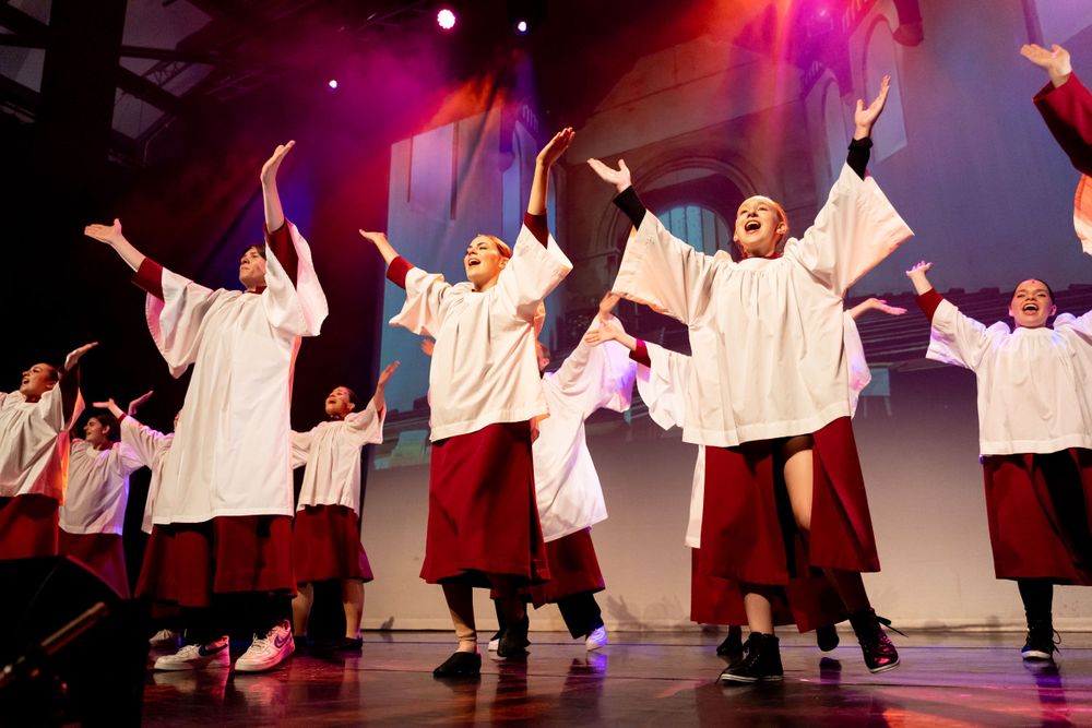 Several performers wearing choir outfits sing a gospel song with their arms outstretched, with projections in the background and pink lights
