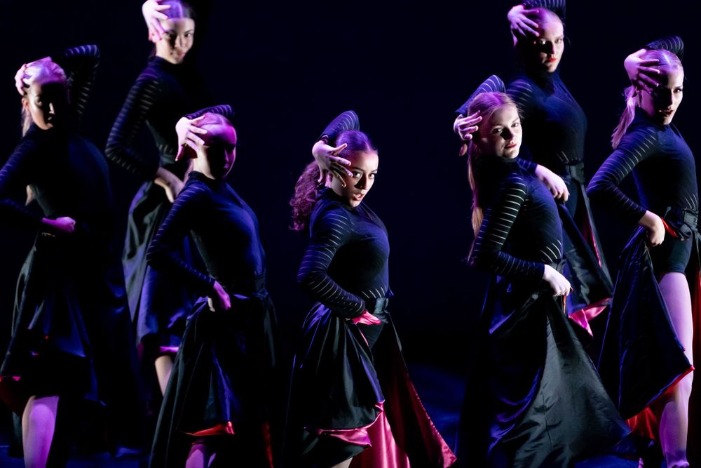 A group of female dancers looking into the camera wearing black tops and pink and black skirts as part of a stage performance