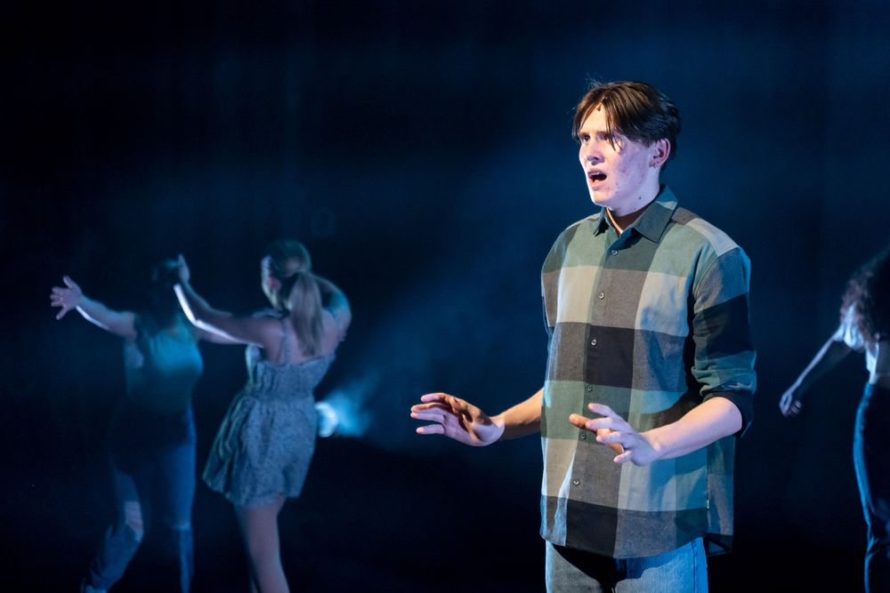 A singer in a spotlight performing a musical theatre song wearing a plaid shirt, against pale blue lighting