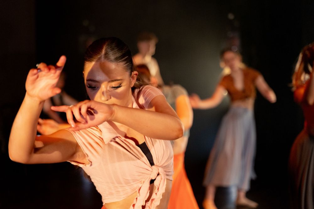Dancer in focus moves expressively onstage, others blurred in background. Dim lighting highlights dynamic motion and expressions.