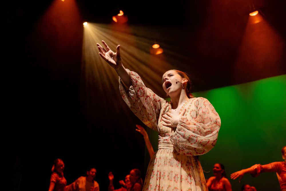 A woman in a patterned dress sings passionately on stage under dramatic lighting, with dancers and a green backdrop behind her.