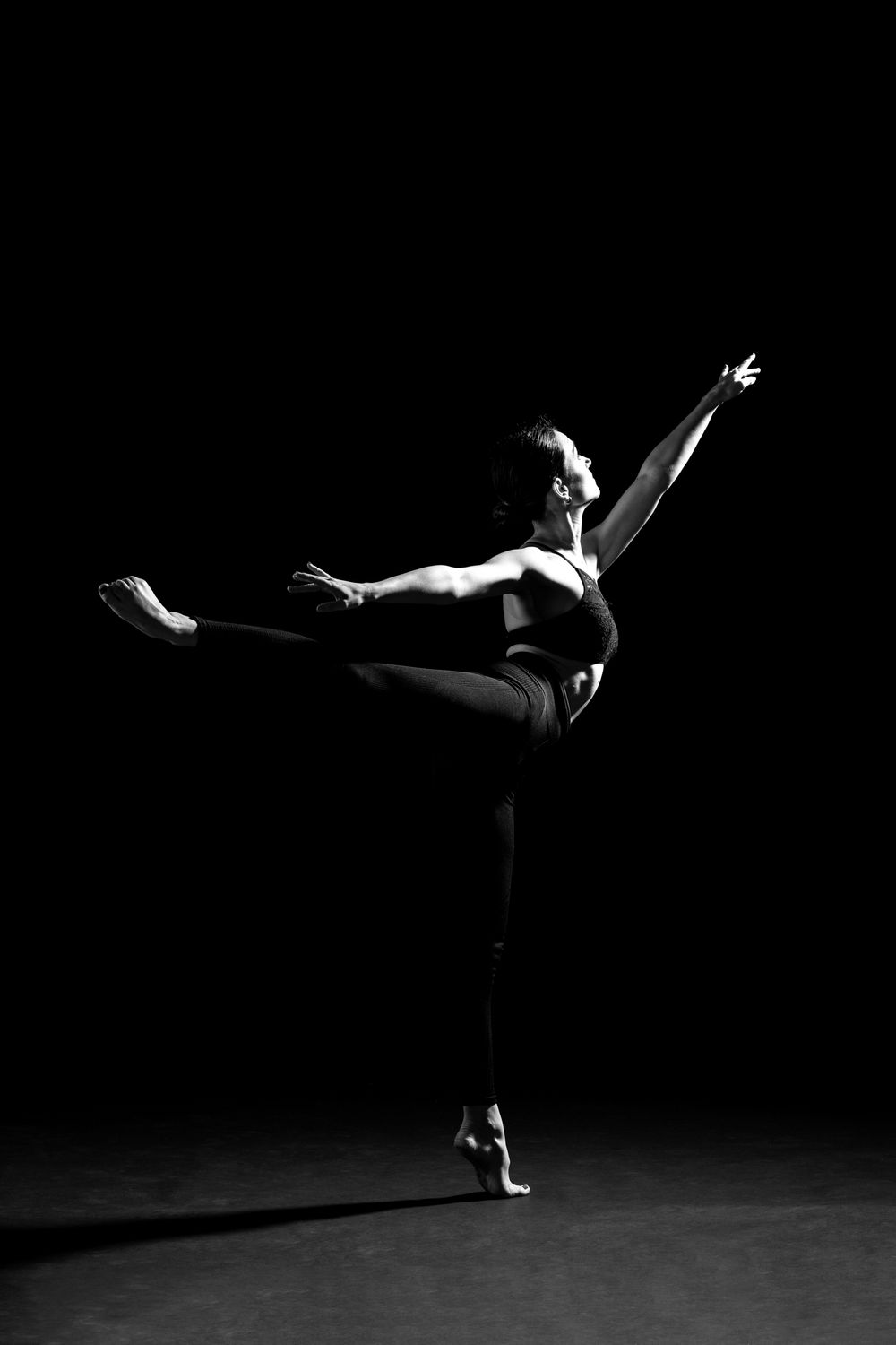 A dancer standing on one foot in arabesque position looking into the light in a moody black and white photograph