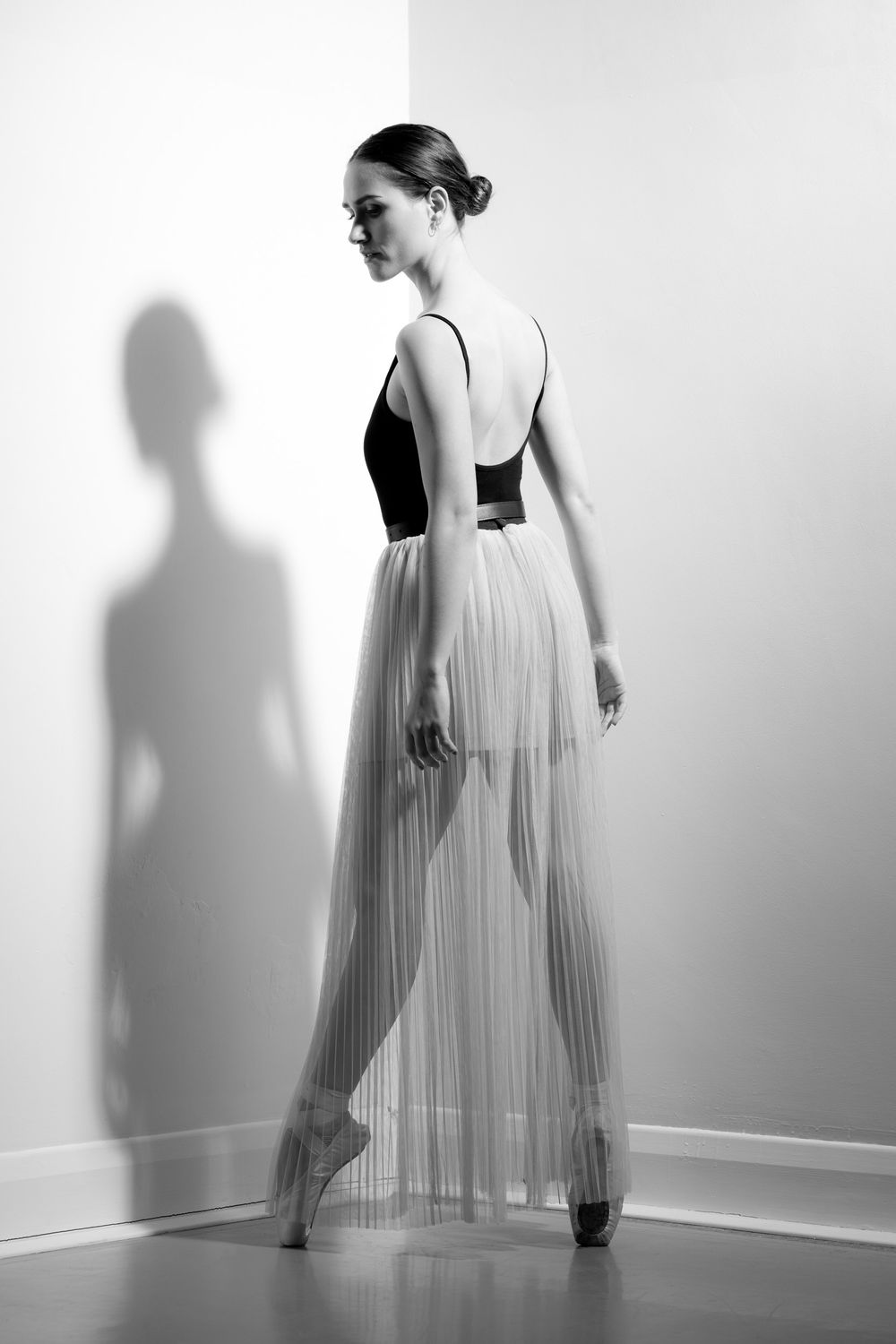 A striking black and white photo of a ballerina en pointe in the corner of a room with high contrast shadow on the wall behind her