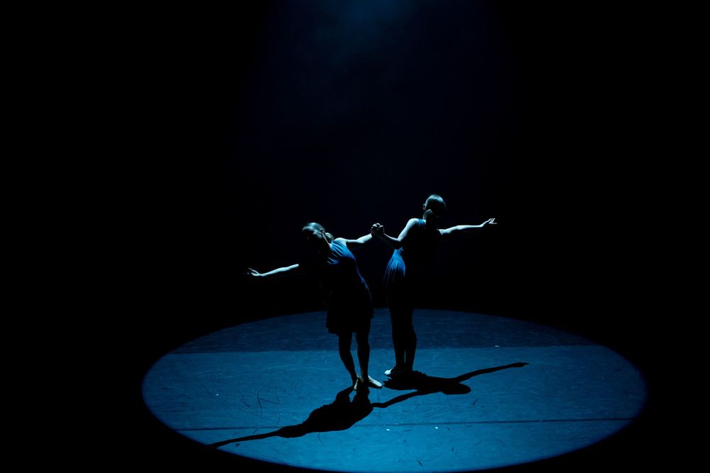 Two dancers perform on a dimly lit stage, casting shadows within a circular spotlight.