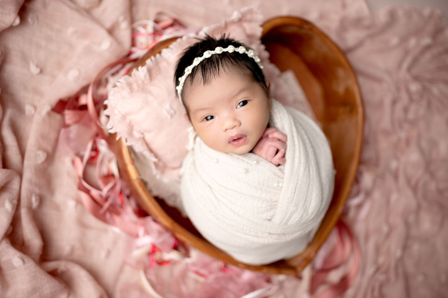 Overhead view of newborn wrapped in white swaddle in wooden bowl with pink backdrop.