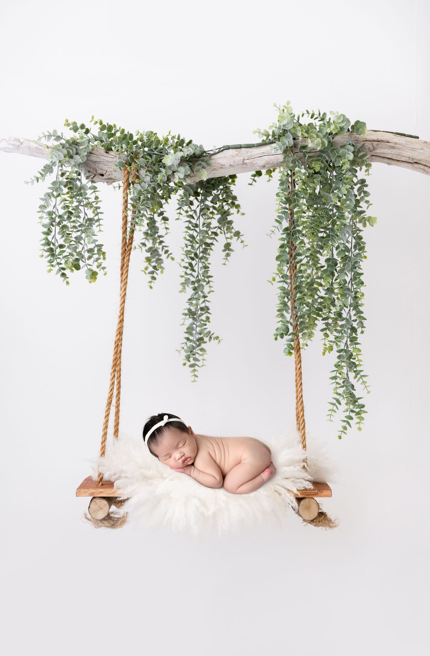 Sleeping newborn on rustic swing decorated with cascading eucalyptus branches.