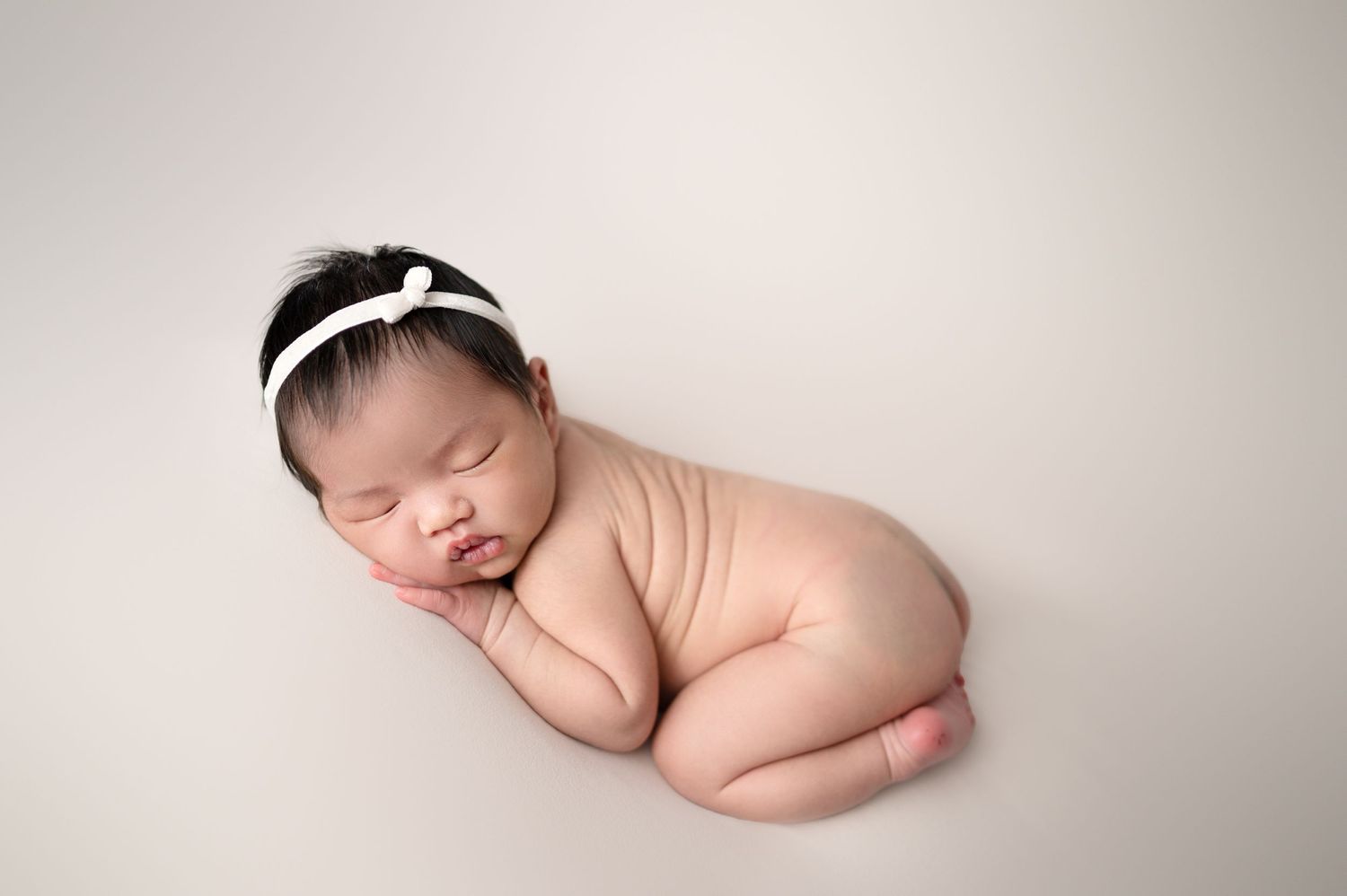 Sleeping newborn with white headband posed in side-lying position on neutral backdrop.