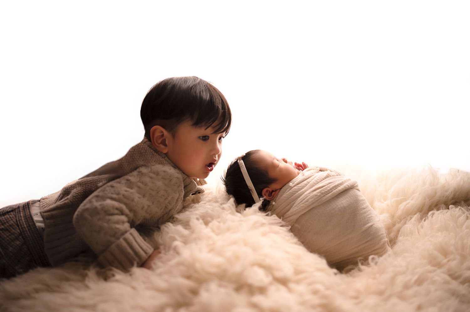 Young child looking at swaddled baby on fluffy white fur backdrop.