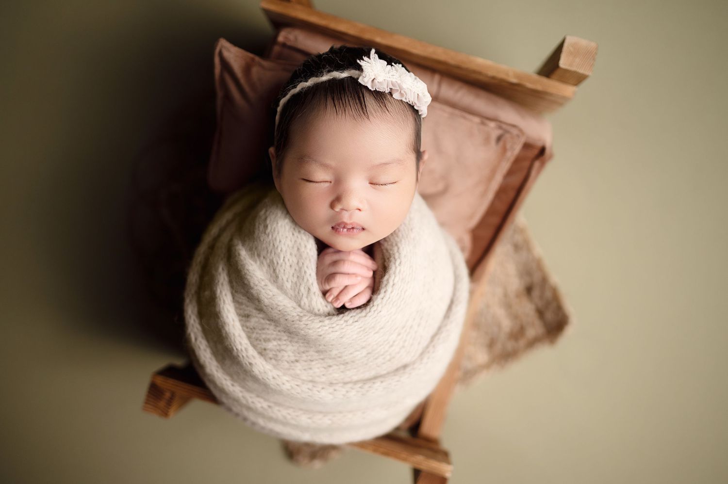 Swaddled sleeping newborn resting in wooden prop with brown draping.