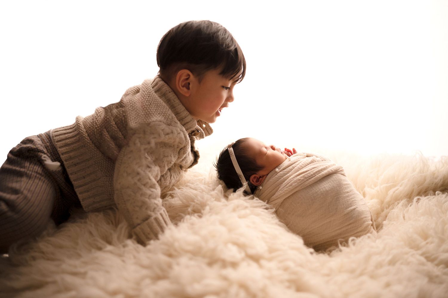 Sibling leaning over to observe swaddled newborn on cream-colored fur surface.