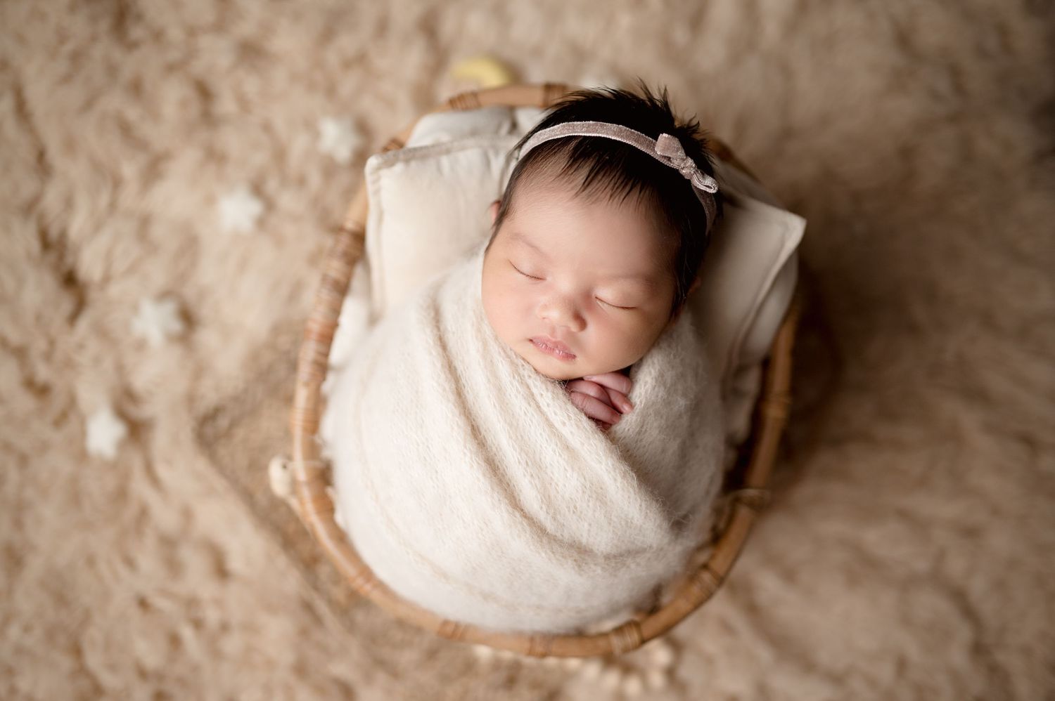 Newborn baby swaddled in white wrap sleeping peacefully in wooden basket on beige backdrop.