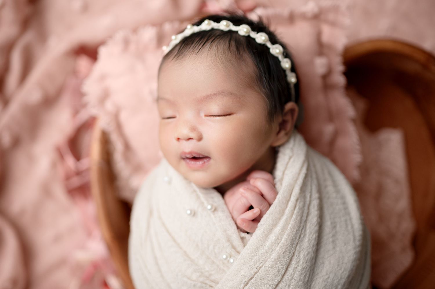 Professional newborn portrait in white wrap with delicate headband against pink background.
