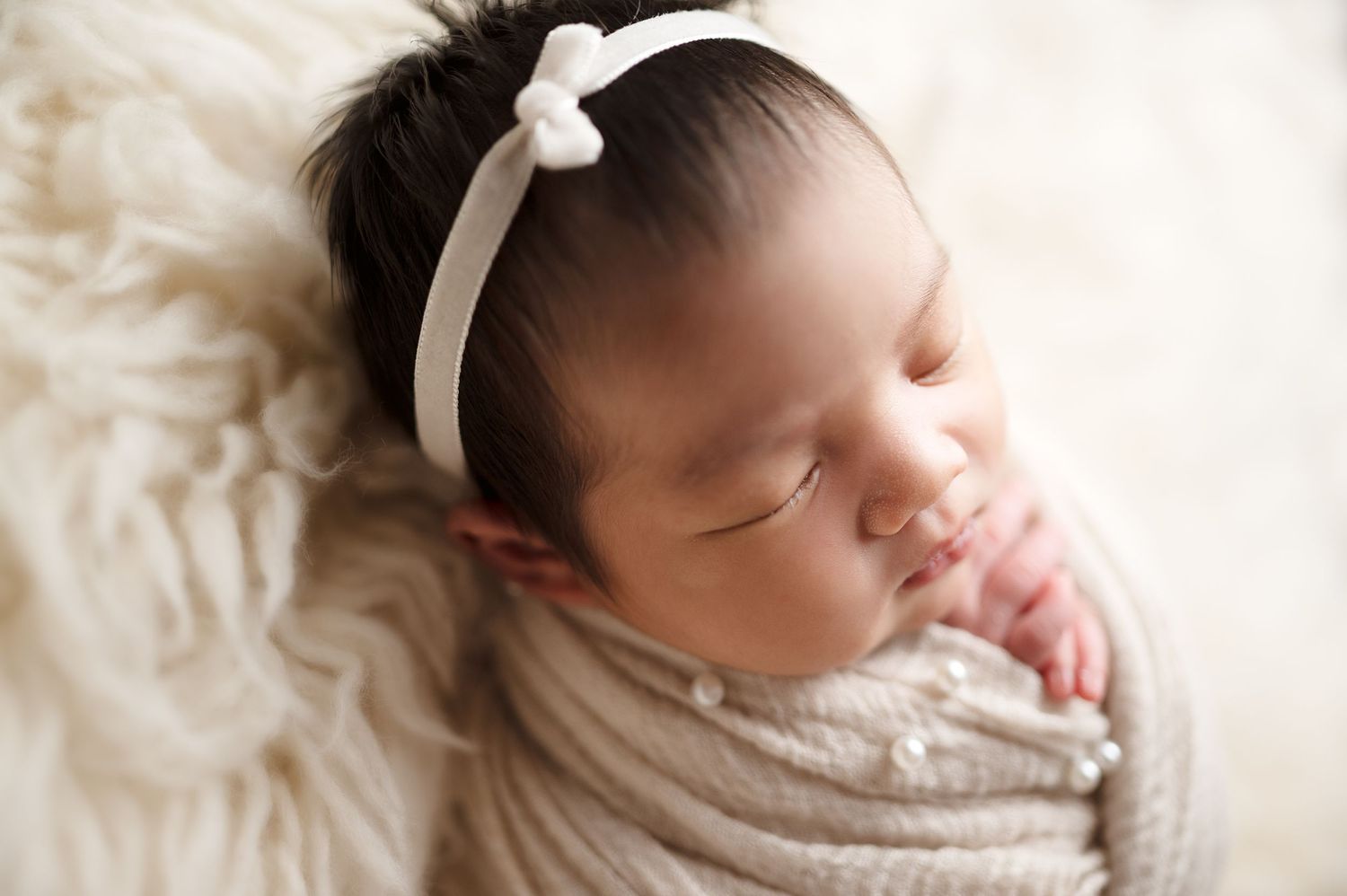 Close-up of sleeping newborn with white headband wrapped in neutral toned fabric.