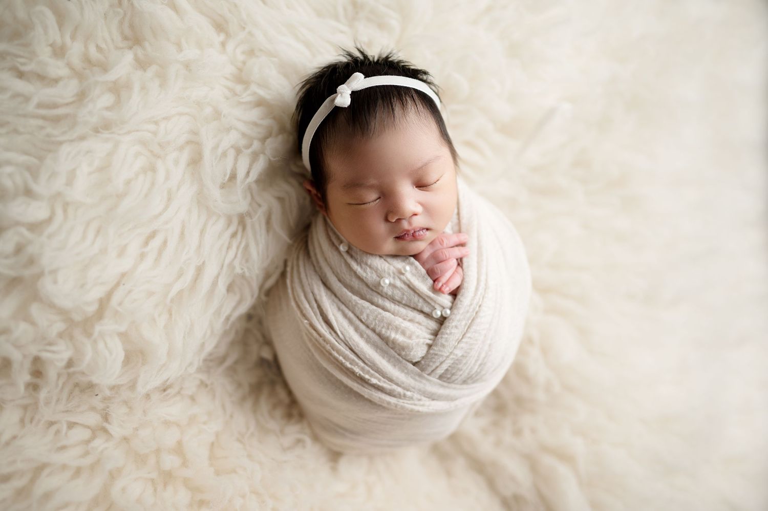 Peaceful newborn wrapped in white fabric sleeping on soft furry white backdrop.
