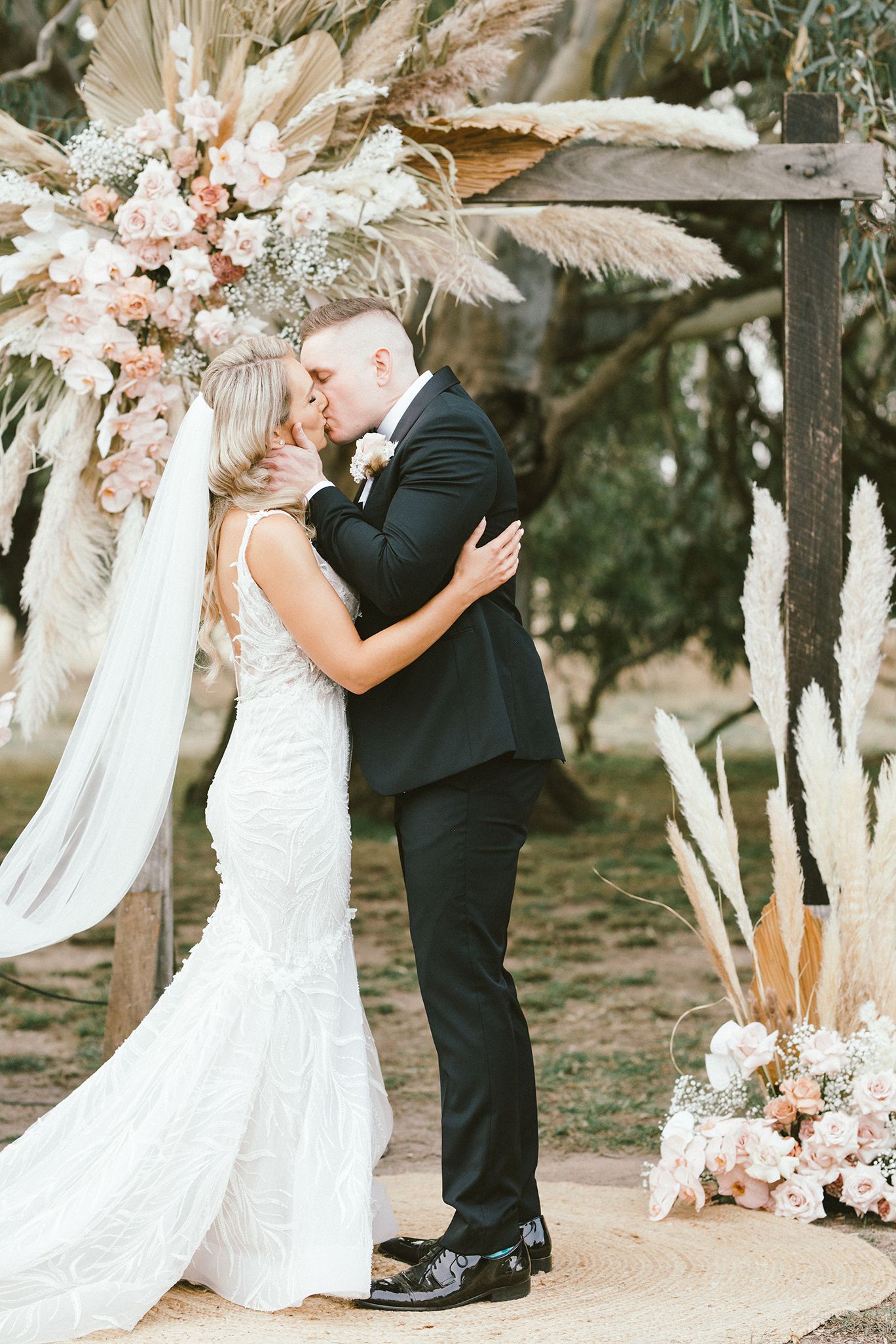 Bride in white lace dress and groom in black suit embrace under floral ceremony arch with dried flowers.