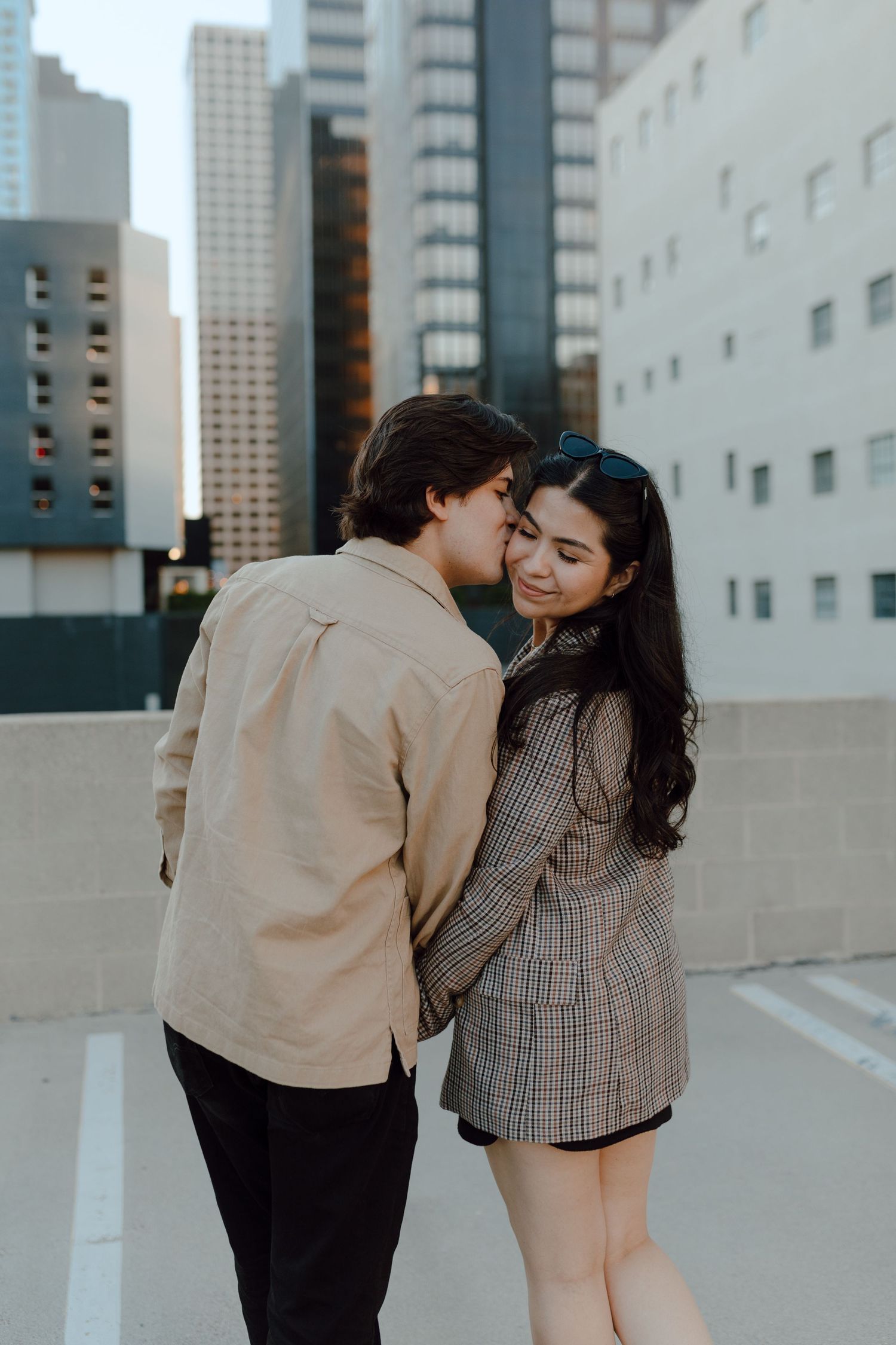 Photograph of couple in the downtown city of Los Angeles. Boyfriend turns to kiss his girlfriend on her temple.