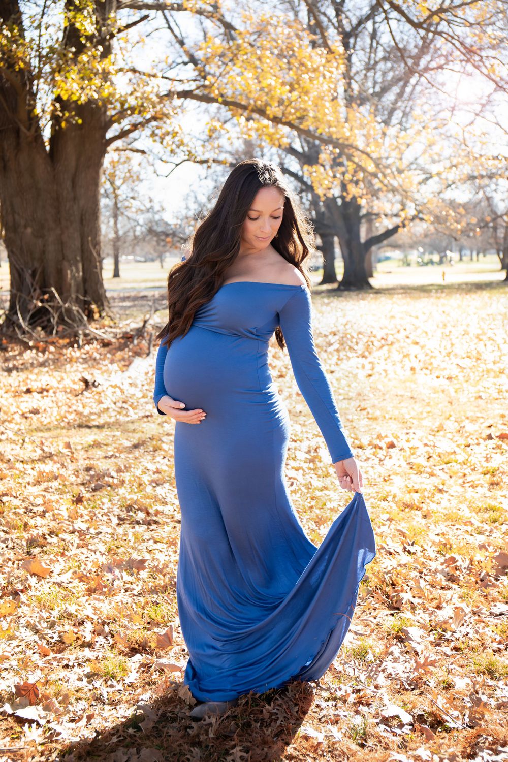 Woman in flowing blue maternity gown poses among falling autumn leaves.