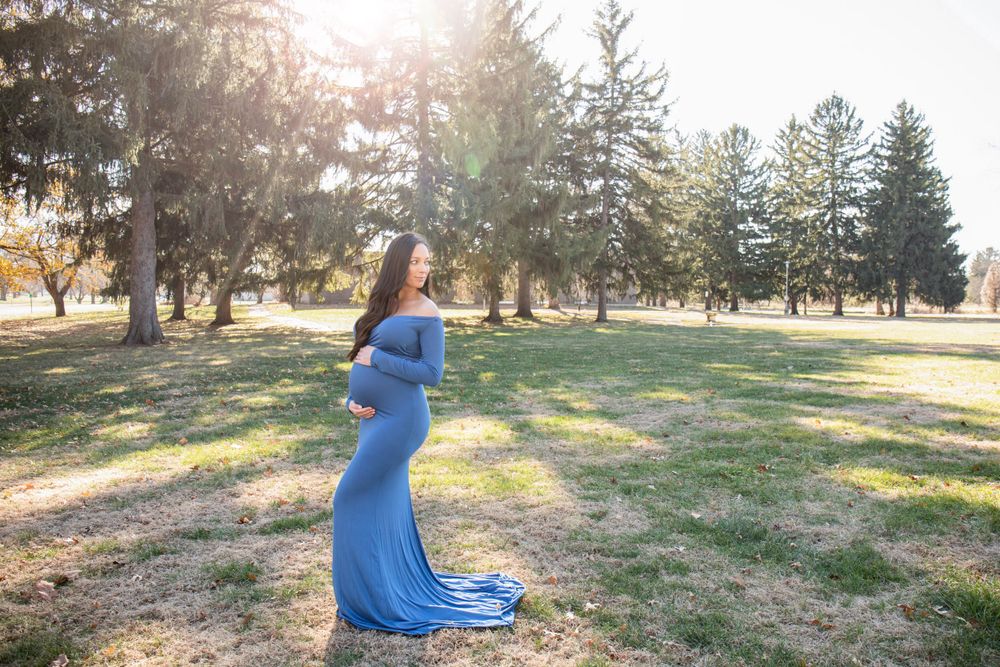 Expectant mother in flowing blue gown poses in sunlit park setting.