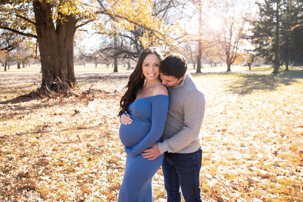 A couple poses for maternity photos in autumn with fallen leaves and warm sunlight.