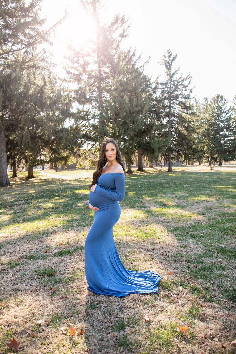 Maternity photo of woman in blue dress with trailing fabric amid pine trees.