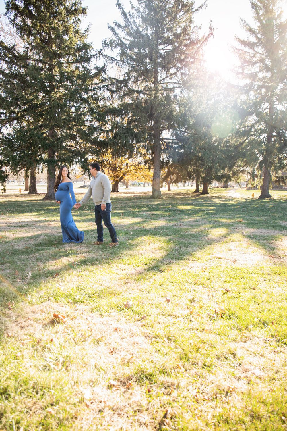 A couple stands in a sunlit park with evergreen trees, creating a romantic atmosphere against golden sunlight.