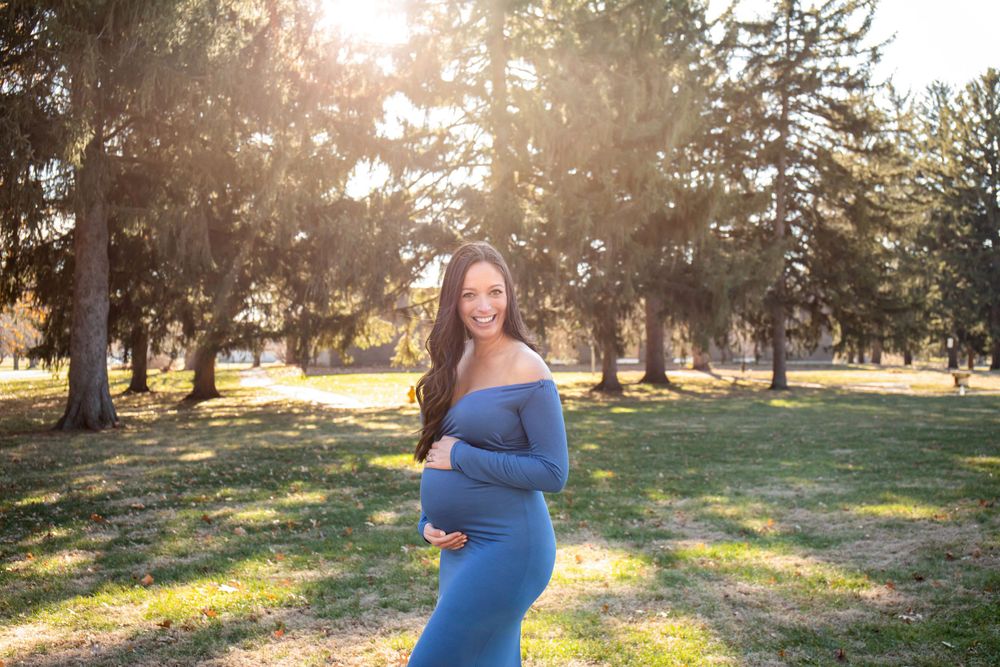 Maternity portrait in blue dress with sunlit evergreen trees in background.