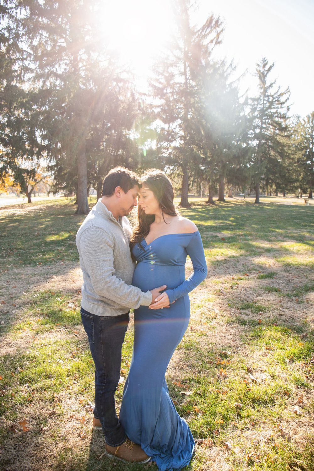 A couple shares an intimate moment in a park with sunlight streaming through evergreen trees.