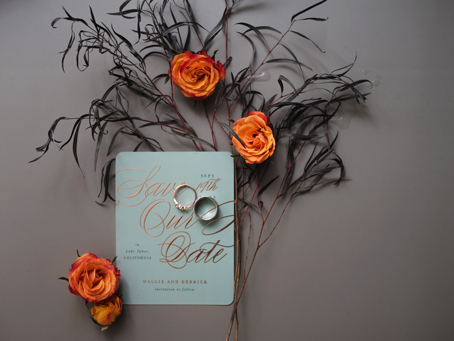 Light blue save the date card with orange roses and dark twigs arranged in a moody flat lay composition.