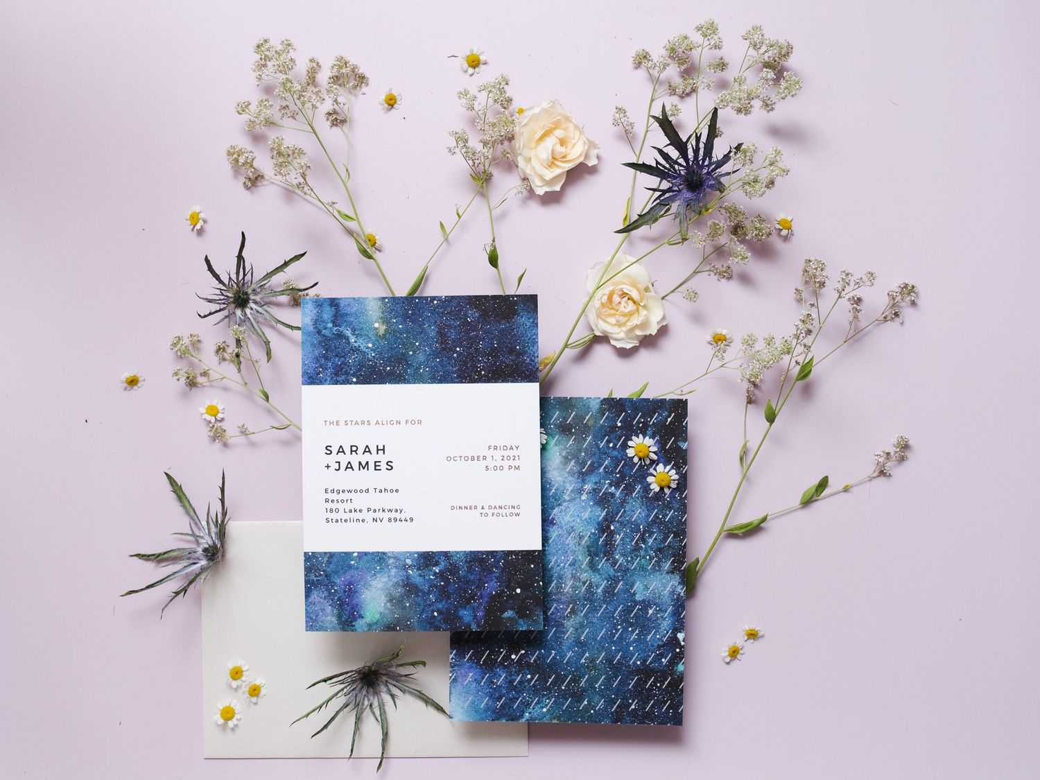 Wedding invitation suite with navy blue watercolor design displayed with delicate white flowers and dried thistles on lilac background.