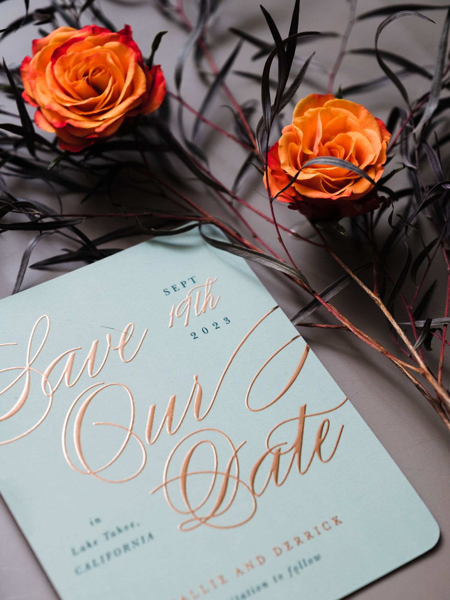 Light blue save the date wedding invitation card with gold calligraphy text and two orange roses on black branches.