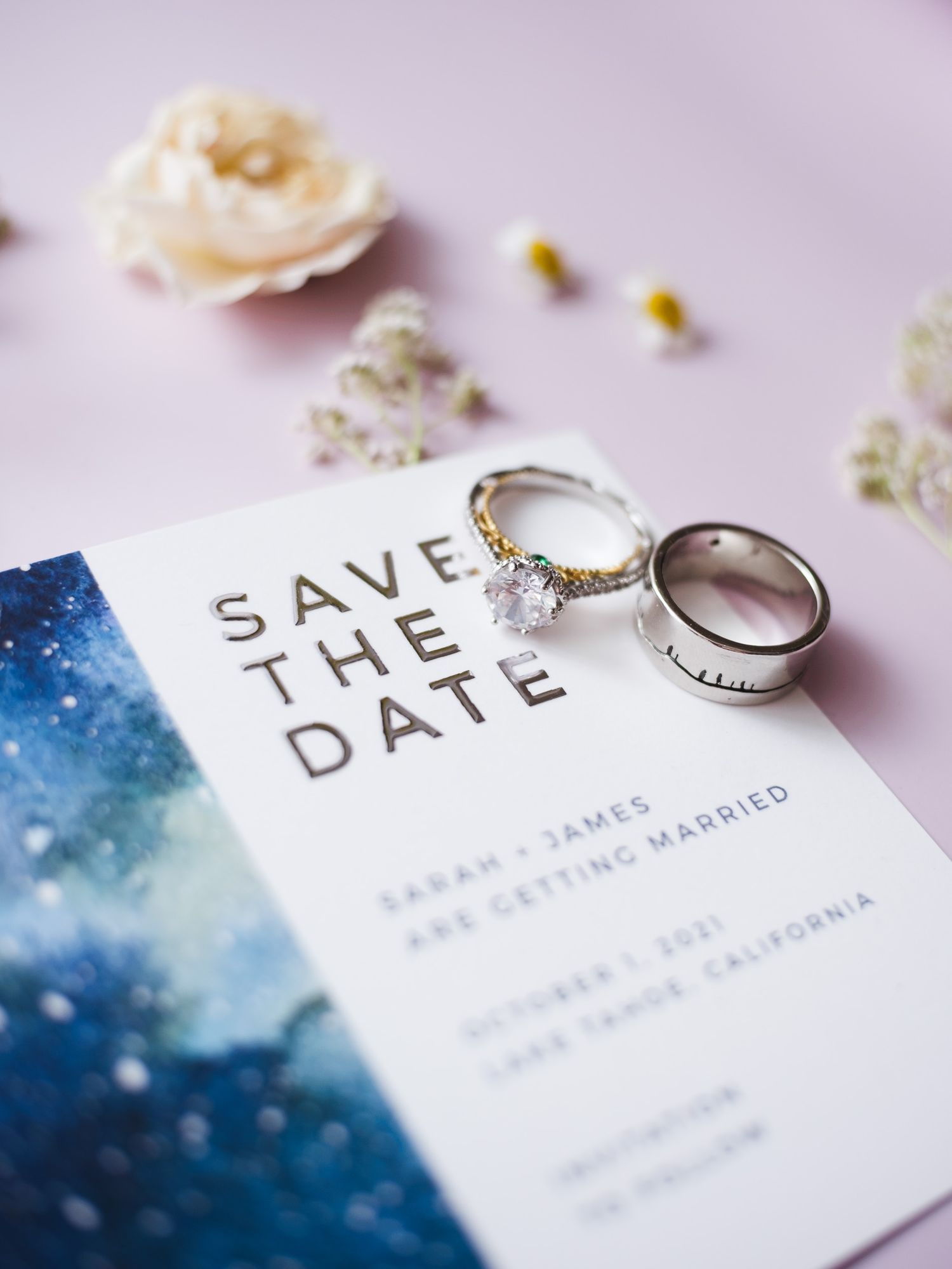 Wedding rings displayed on a save the date invitation card with watercolor blue design and a small white rose accent.