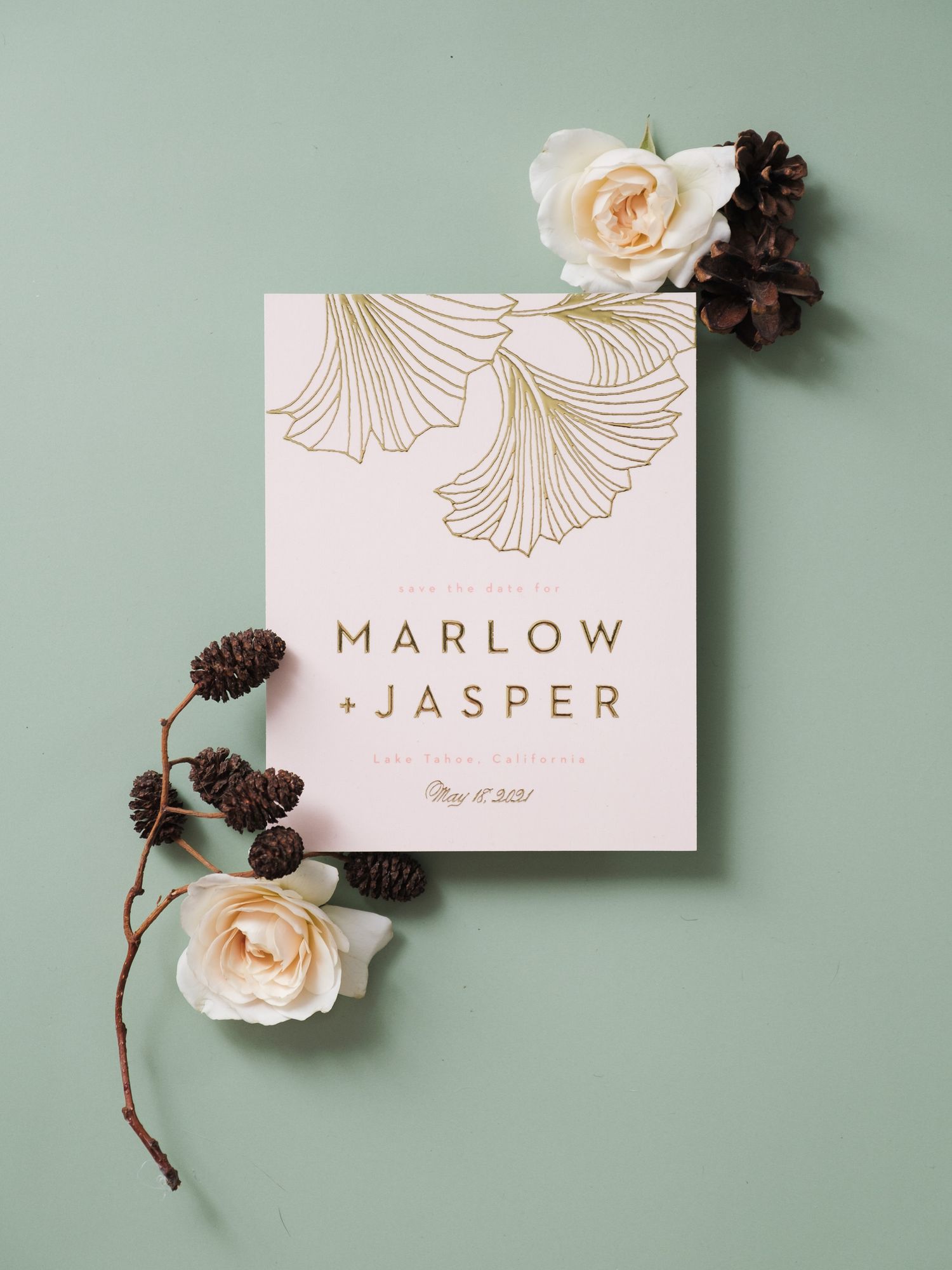 Elegant blush wedding invitation with gold ginkgo leaf design adorned with peach roses and pinecones on sage background.