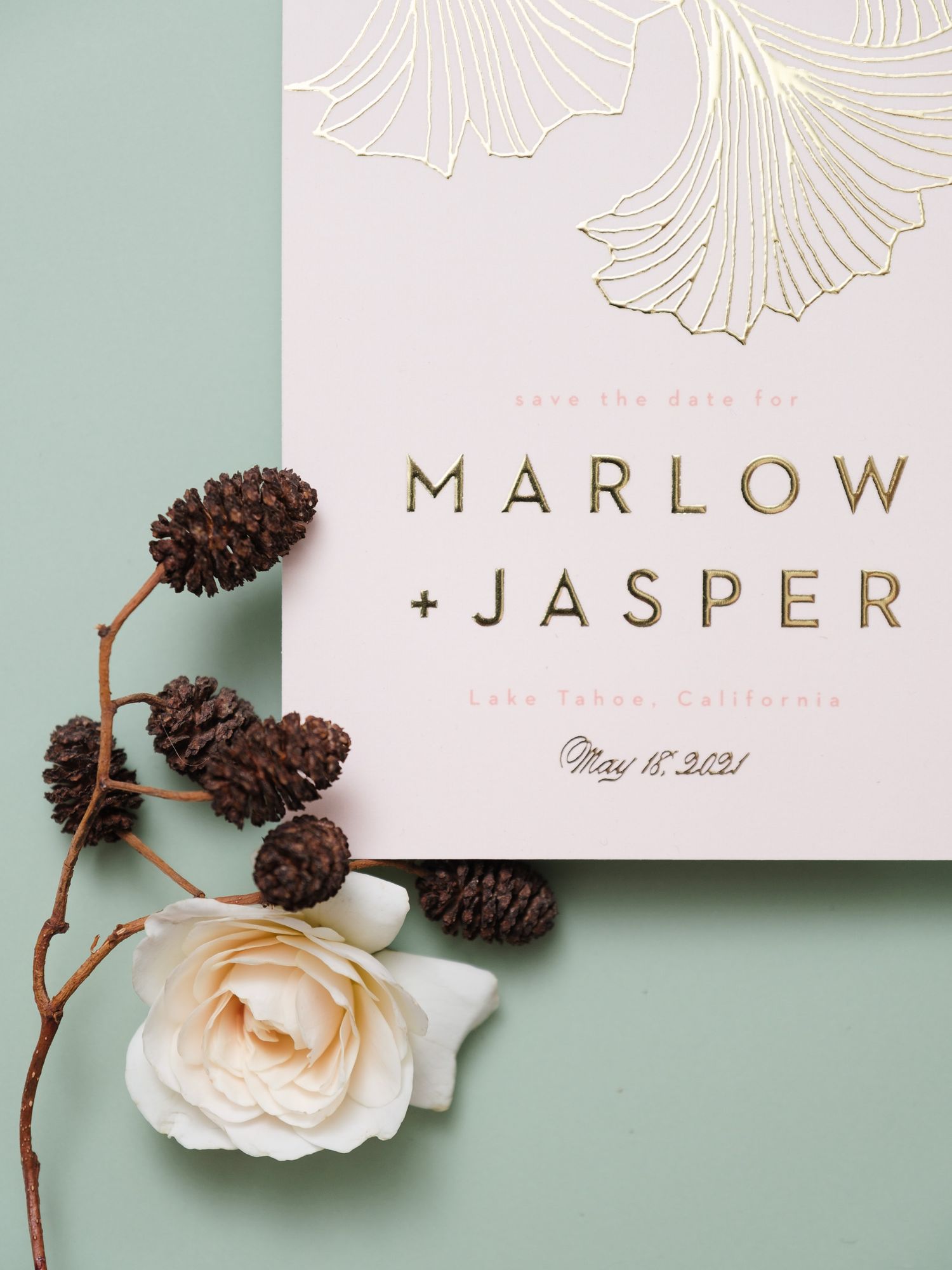 Wedding invitation with gold foil ginkgo leaves, pinecones and a white rose on sage green background.