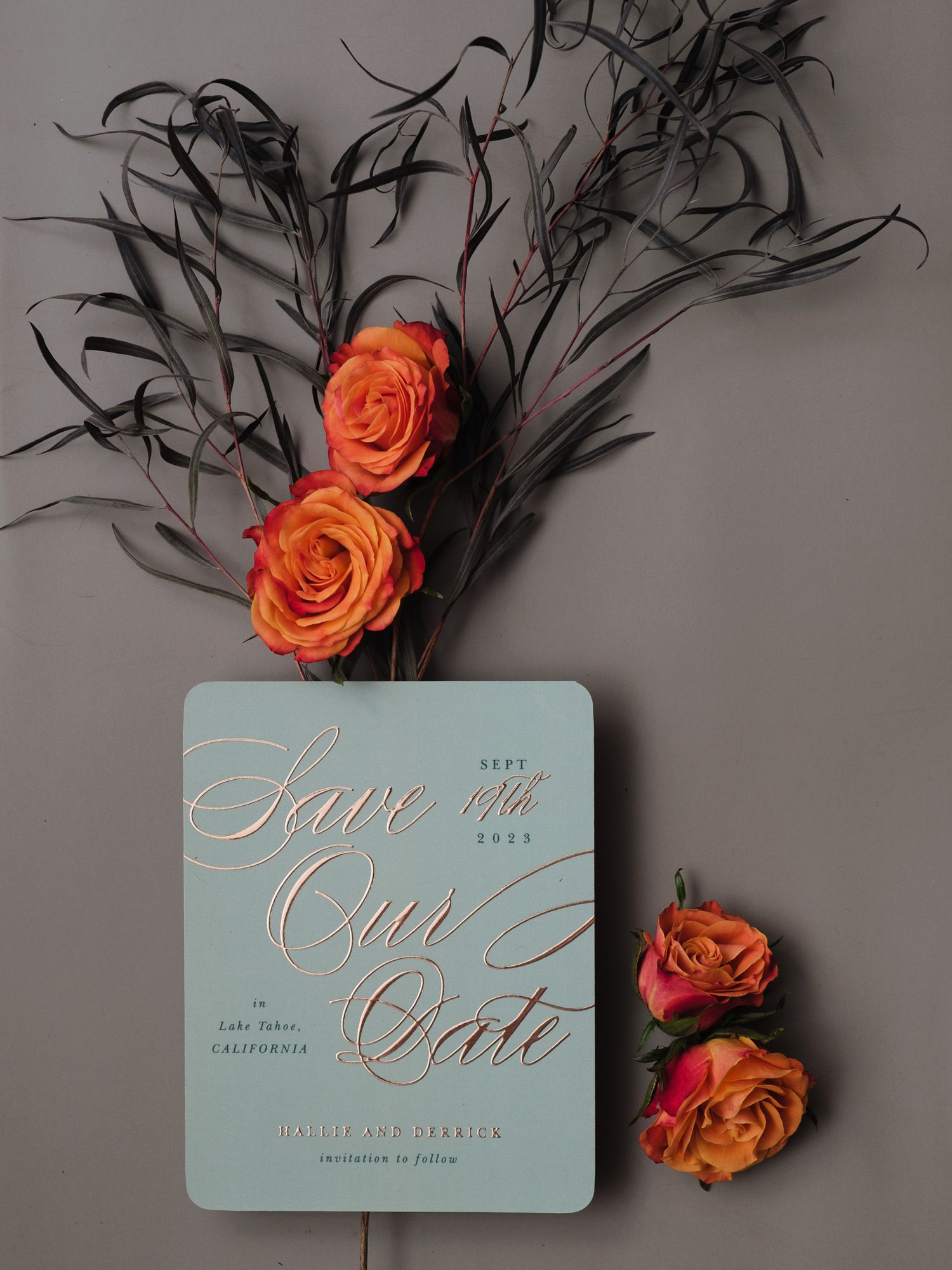 Elegant mint green save-the-date invitation card with orange roses and black feathers against a gray background.