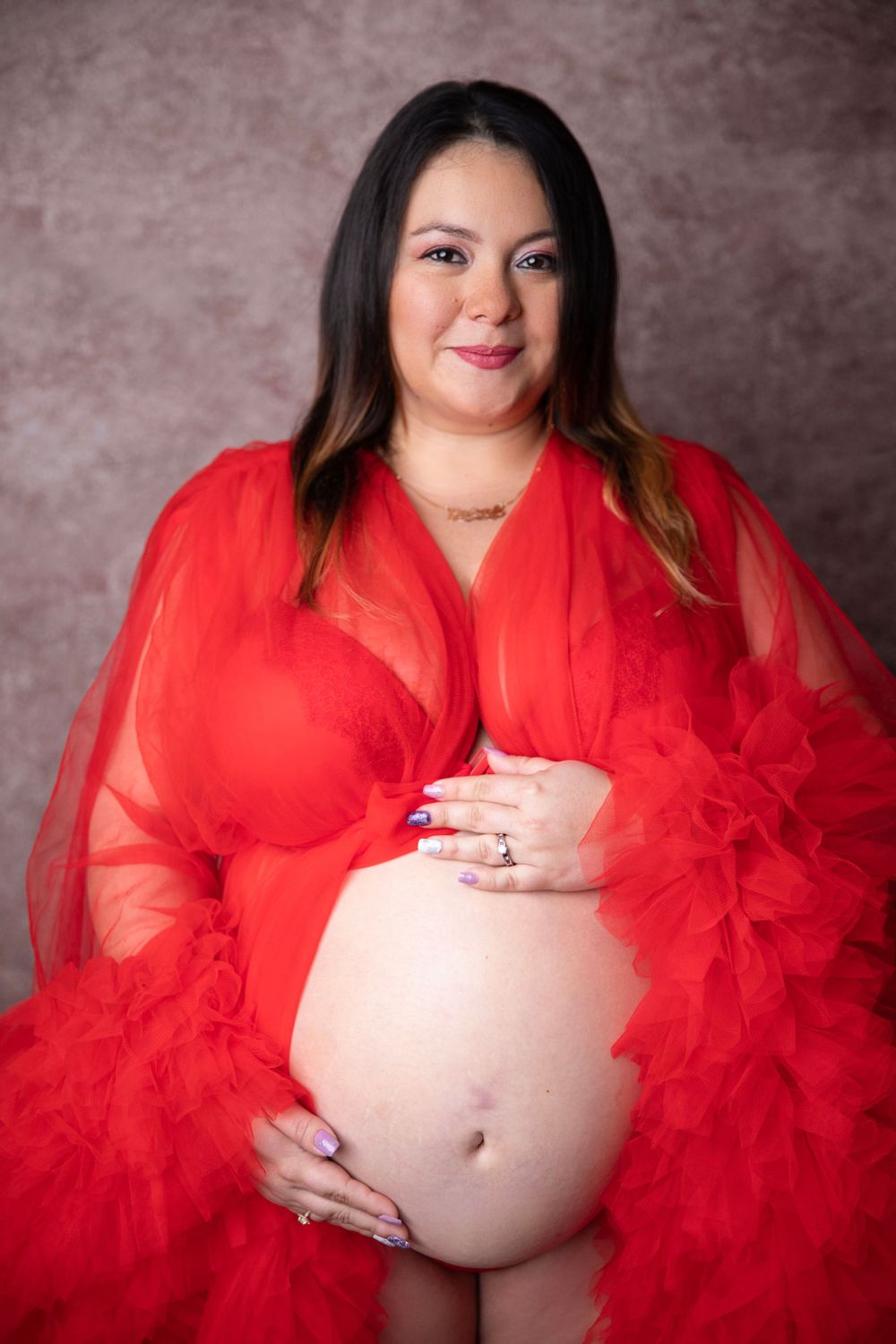 Pregnant woman in flowing red tulle robe showcasing bare baby bump in studio maternity portrait.