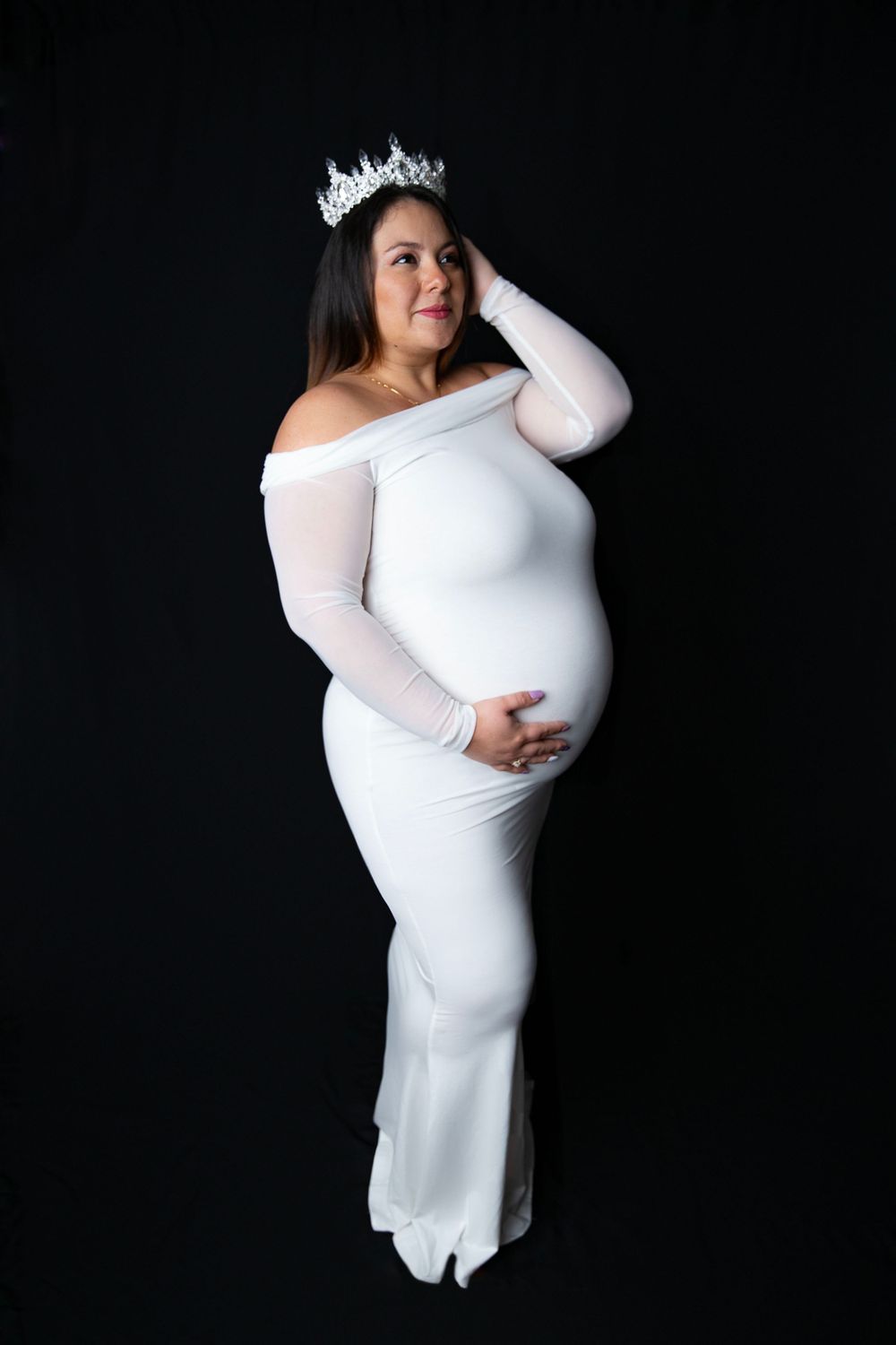 Elegant pregnant woman in white off-shoulder fitted gown wearing a crystal crown against black background.