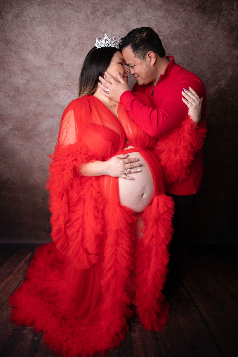 Romantic maternity portrait of couple in red outfits, forehead to forehead, hands on pregnant belly.