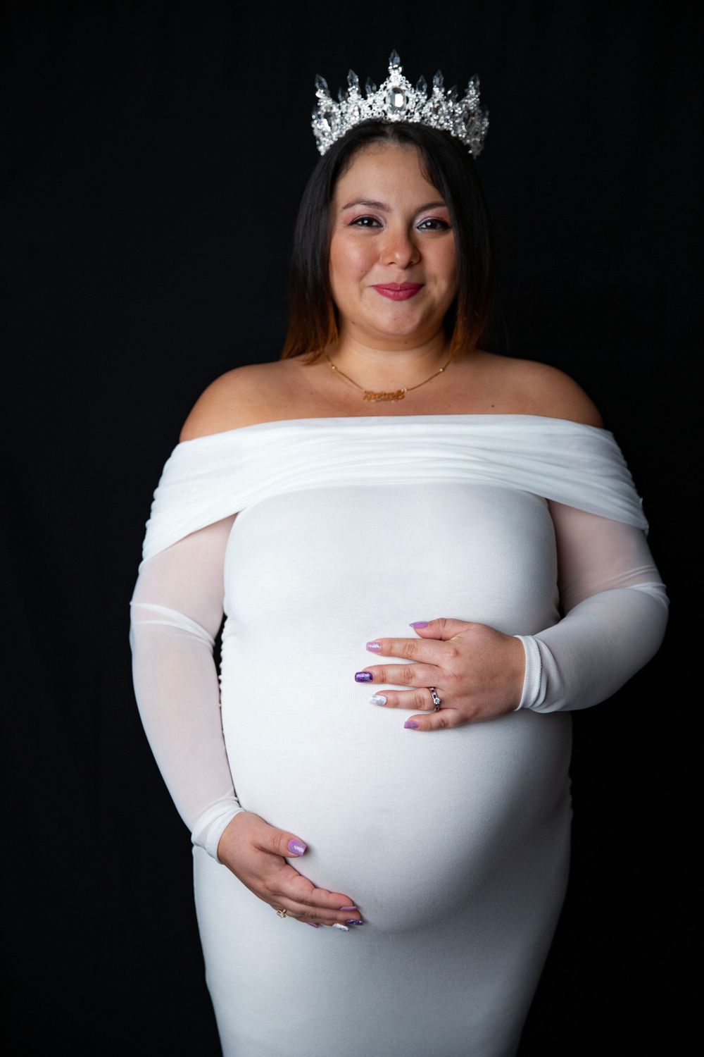 Smiling pregnant woman in white off-shoulder maternity dress with crystal tiara cradling baby bump.