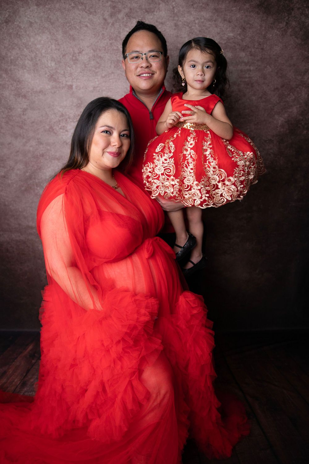 Family maternity portrait with pregnant woman in red tulle gown, man in red shirt, and young girl in red dress.
