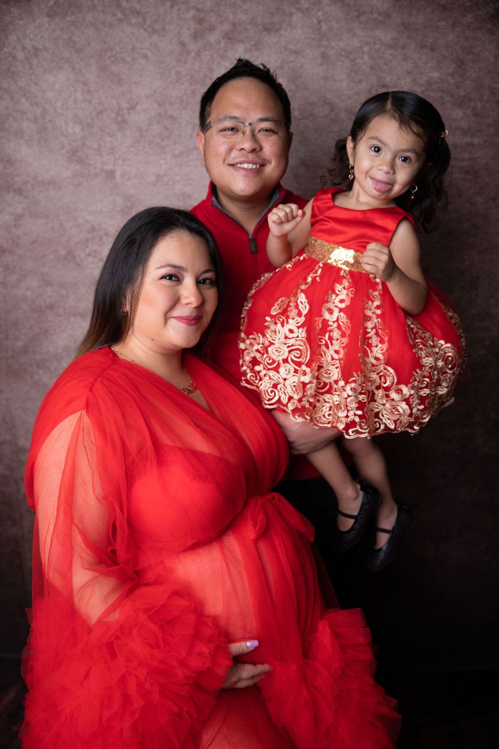 Joyful maternity photo of pregnant woman in red tulle gown with husband and young daughter in matching red dress.
