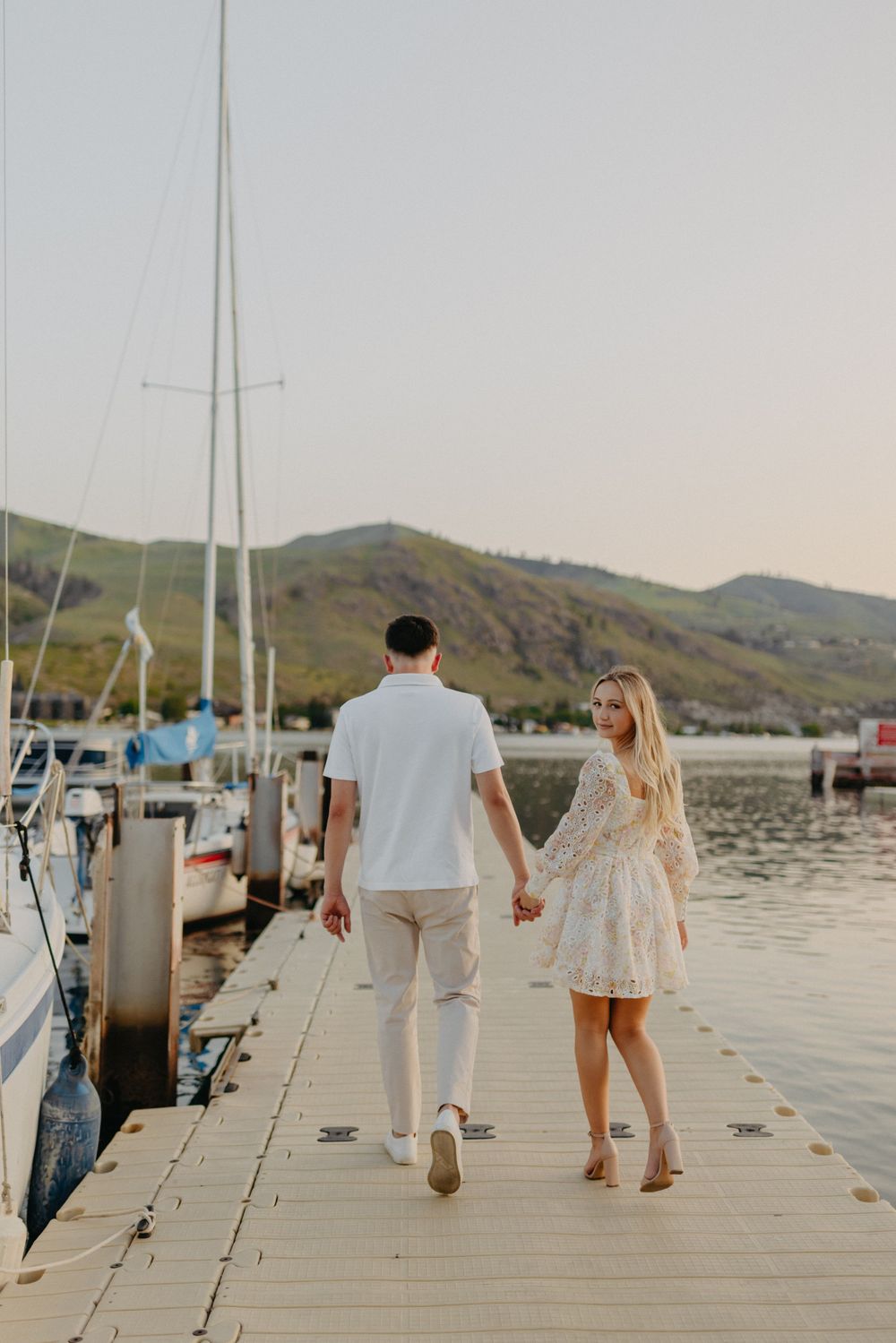 A couple walks hand in hand along a marina dock with sailboats and mountains in view.