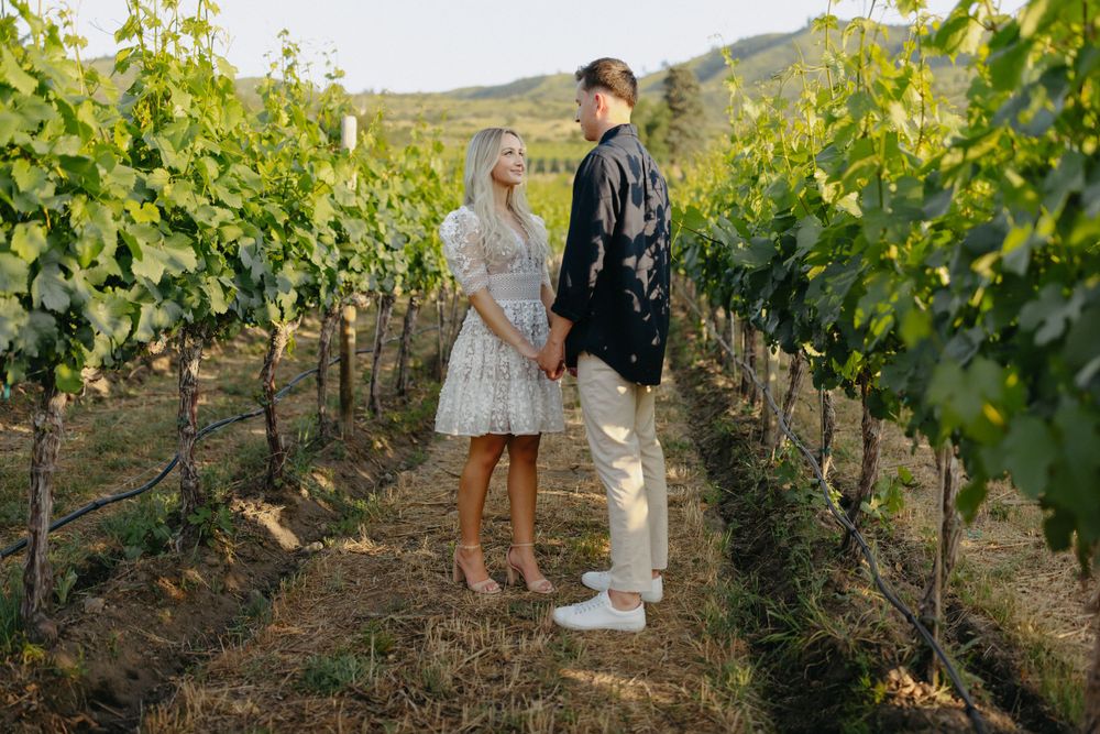 A couple stands between rows of grapevines during a sunny afternoon.