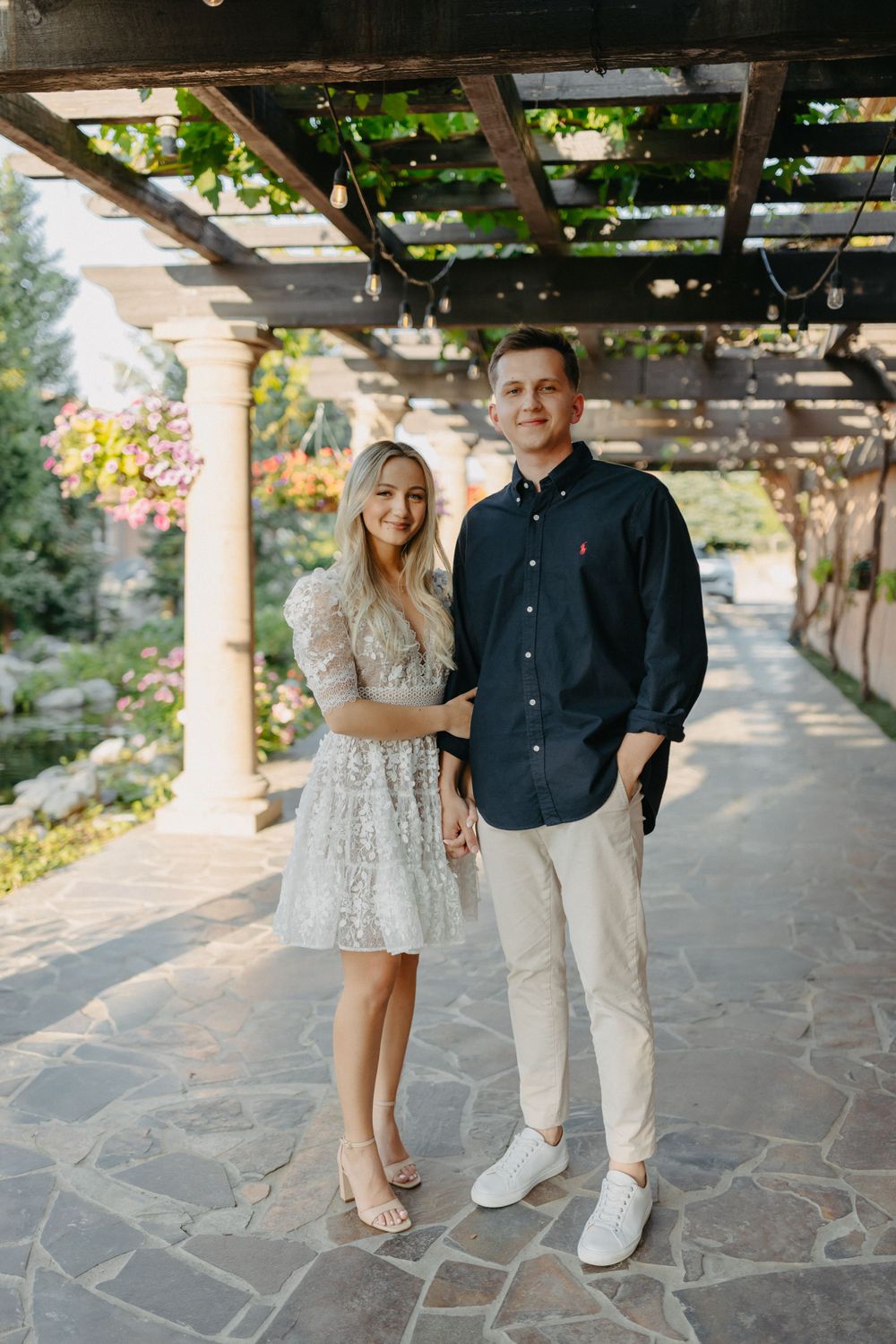 A couple poses together under a pergola in formal attire on stone pavers.