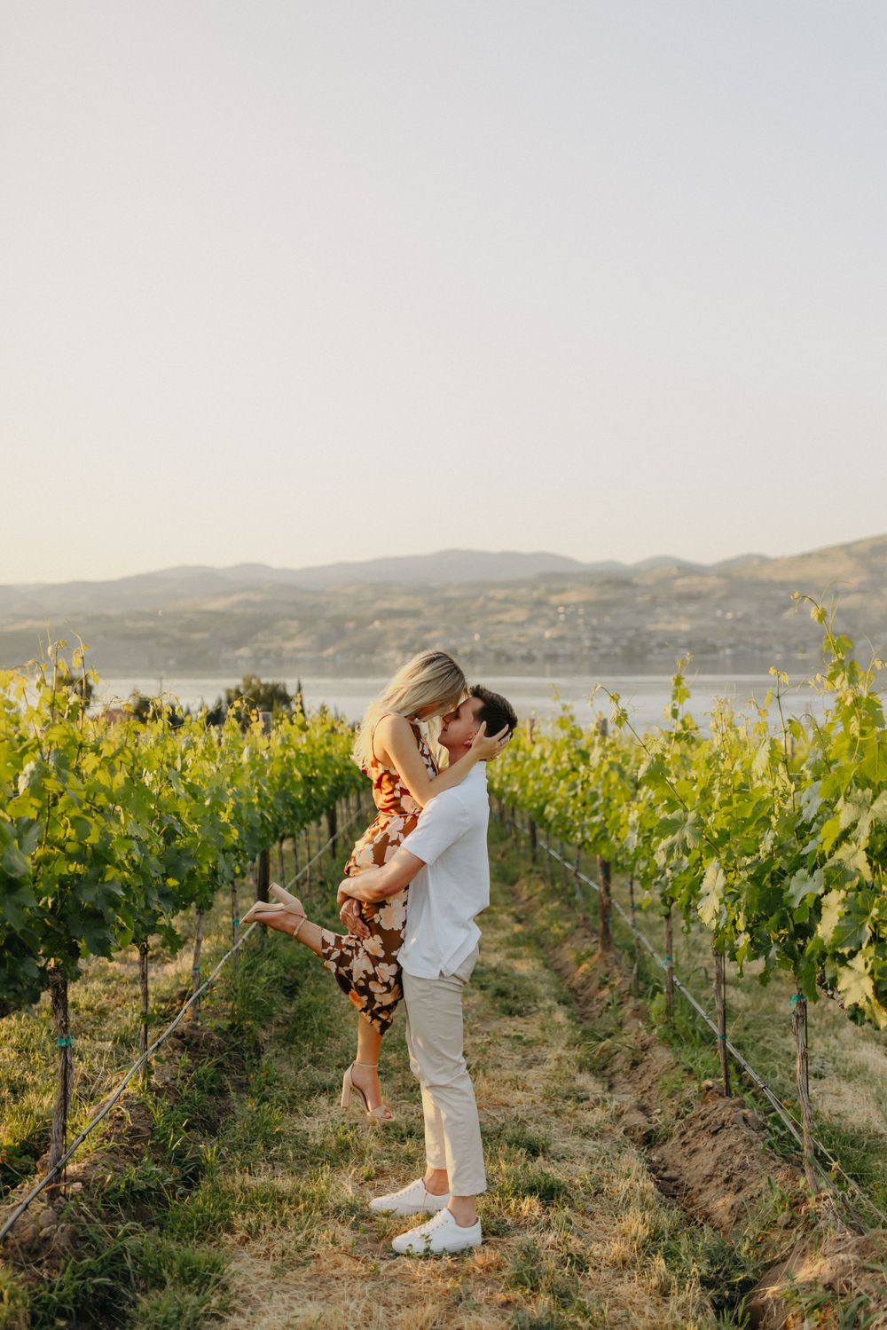 A couple sharing a romantic moment in a vineyard with a lake and mountains in the background.
