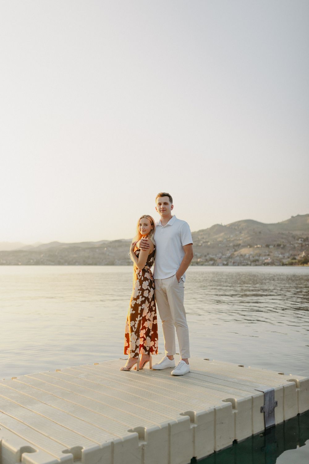 A couple standing together on a dock by the lake during sunset wearing casual summer attire.