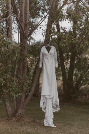 White lace wedding dress hanging on a tree branch in a rustic outdoor setting.