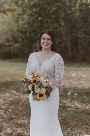Bride holding sunflower bouquet poses in lace wedding dress outdoors.
