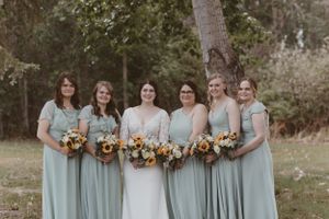 Bridal party in mint green dresses posing with sunflower bouquets in a forest setting.