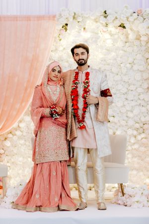 Traditional South Asian wedding couple in pink and cream formal attire against floral backdrop.